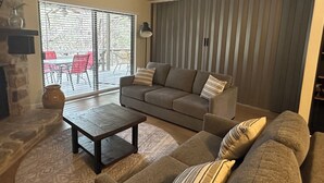 A living room with a sofa, a coffee table, and a fireplace. There is a sliding glass door leading to an outdoor area with chairs.