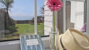 A window with a view of a garden and the sea, a pink flower in a vase, a framed book, and a straw hat on a table. - Watchtower (Torpoint)