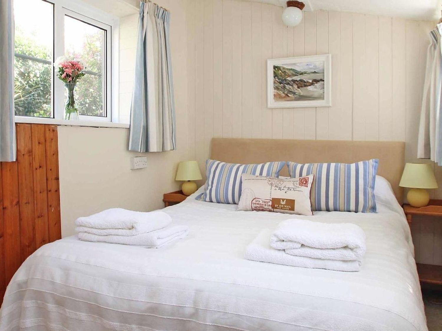 A neatly made bed with white linens and striped pillows, a bedside table with a lamp, a window with curtains, and a painting on the wall.