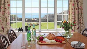 Una mesa de comedor con vistas al mar a través de amplios ventanales, una botella de agua, una botella de vino, una hogaza de pan, queso, tomates y un jarrón con flores.