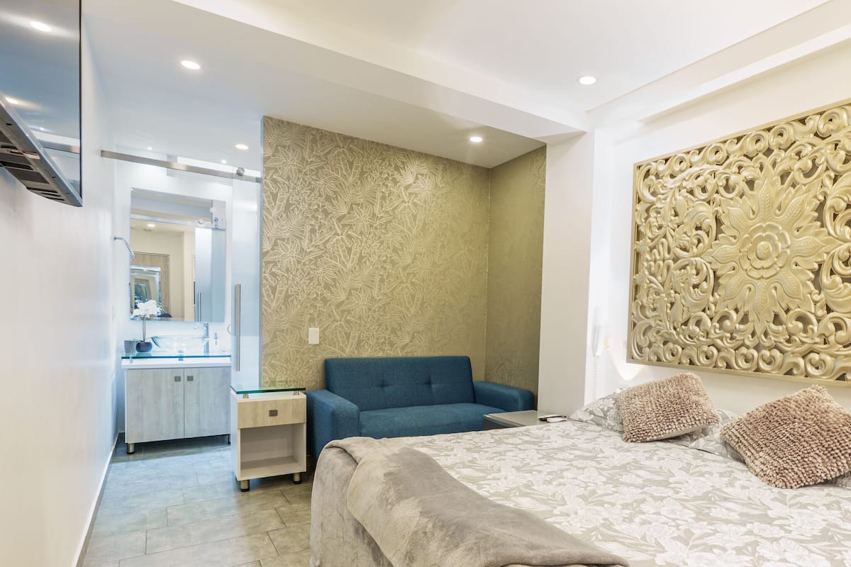 Quarto moderno com cama, sofá azul, mesa de cabeceira e painel decorativo na parede.