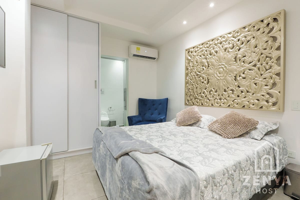 Quarto com cama, uma cadeira azul, guarda-roupa e painel decorativo na parede.