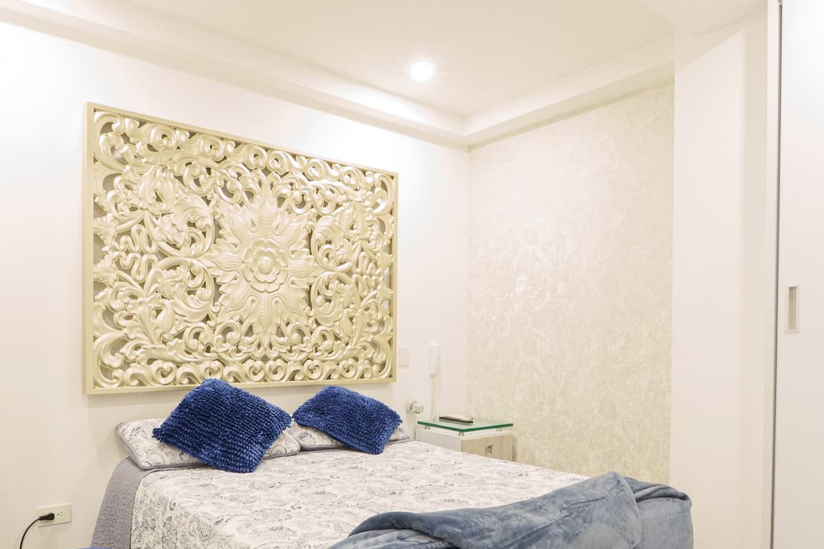 Quarto com cama, um banco azul, painel decorativo na parede e mesa de cabeceira com abajur.