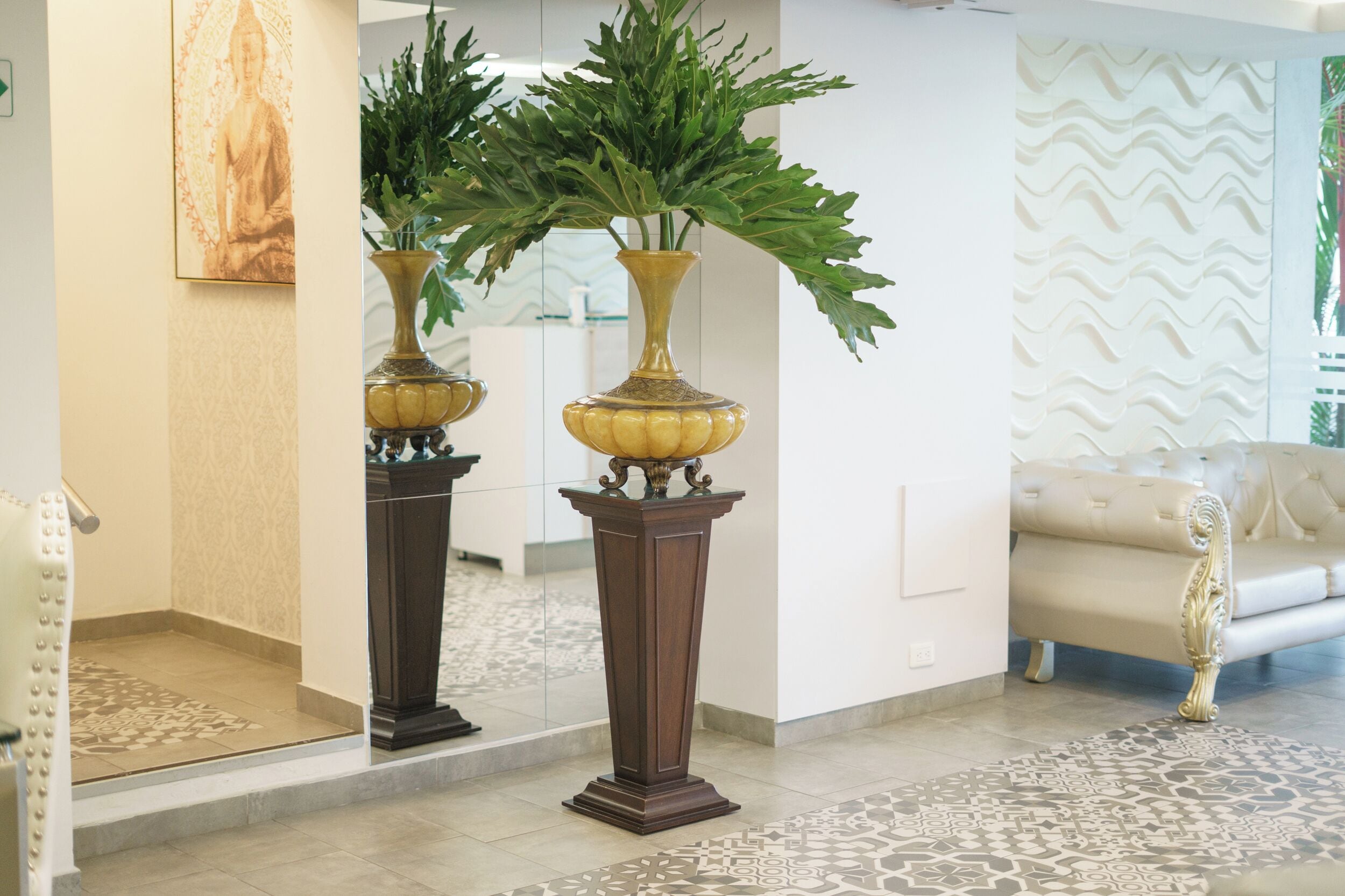 A reception area with a large mirror, a Buddha statue, two potted plants, a patterned rug, and a white sofa.