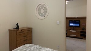 A bedroom with a bed, a wooden dresser, a TV on a wooden cabinet, and a wall-mounted window. - Centro da Cidade 2 quartos cama de casal (Cabo Frio)