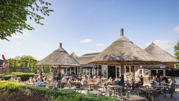 Ein Straßencafé mit strohgedeckten Tischen und Stühlen, umgeben von viel Grün.
