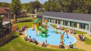 Ein Spielplatz mit Rutschen und Pool, umzäunt und mit einem Gebäude mit großen Fenstern.