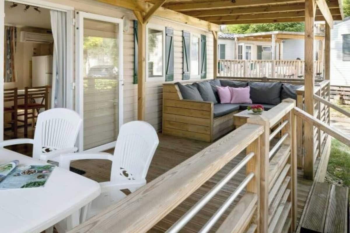 Un espace extérieur abrité, doté d’une terrasse en bois, d’un canapé avec des coussins et de chaises en plastique blanches.