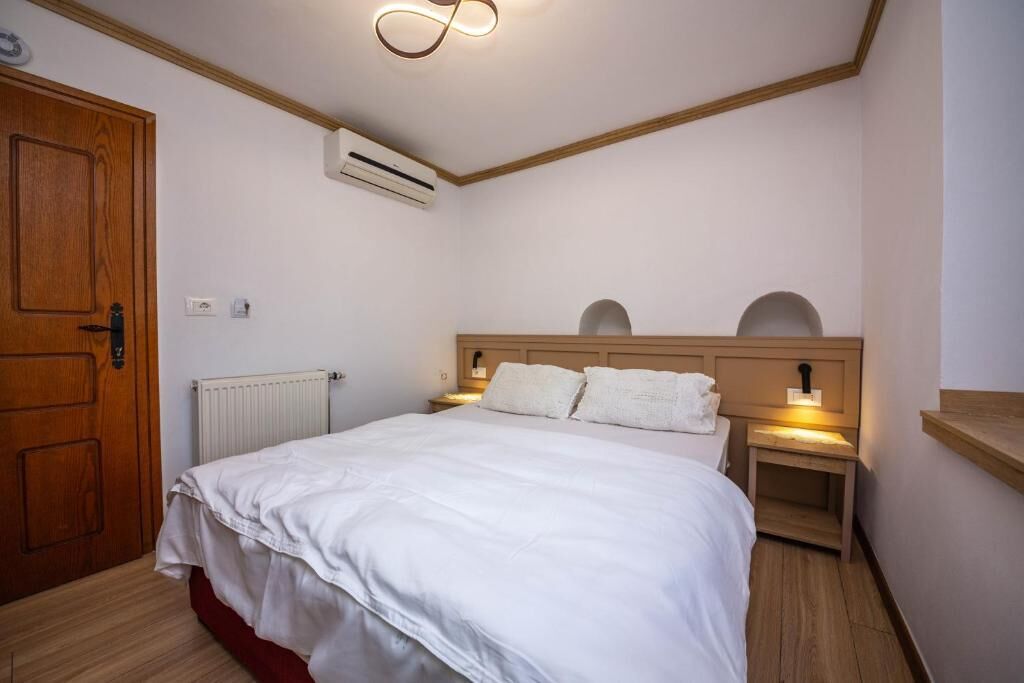 Une chambre avec une porte en bois, du linge de lit blanc, une tête de lit agrémentée de deux tables de chevet et un appareil de climatisation fixé au mur.