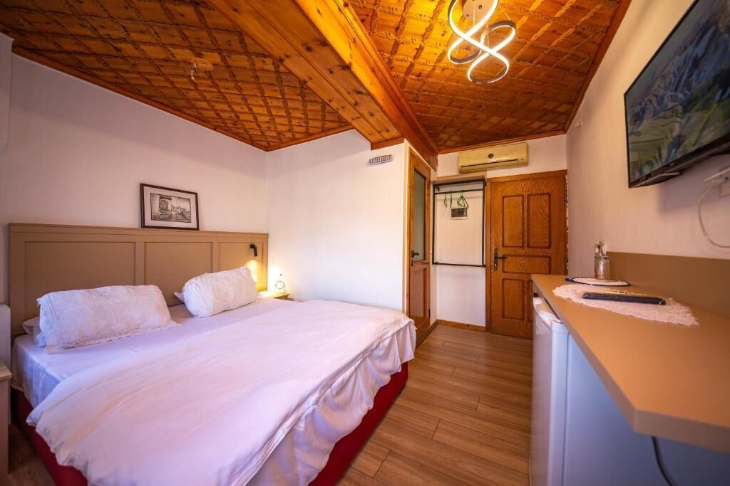 A hotel room with a wooden ceiling, a bed with white linens, a small desk with a lamp, and a television on the wall.