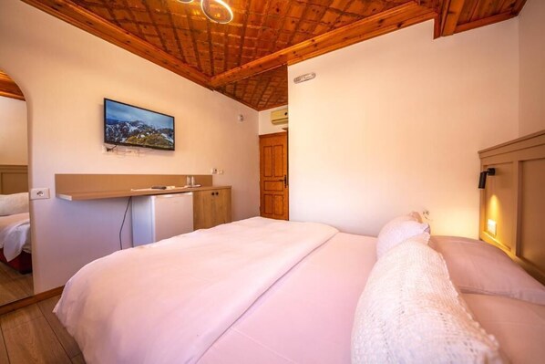 Une chambre avec un plafond en bois, un lit avec une literie blanche, un petit bureau et un téléviseur fixé au mur.