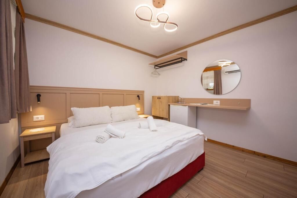 A hotel room with a large bed, a bedside table, a mirror, and a small desk with a lamp.