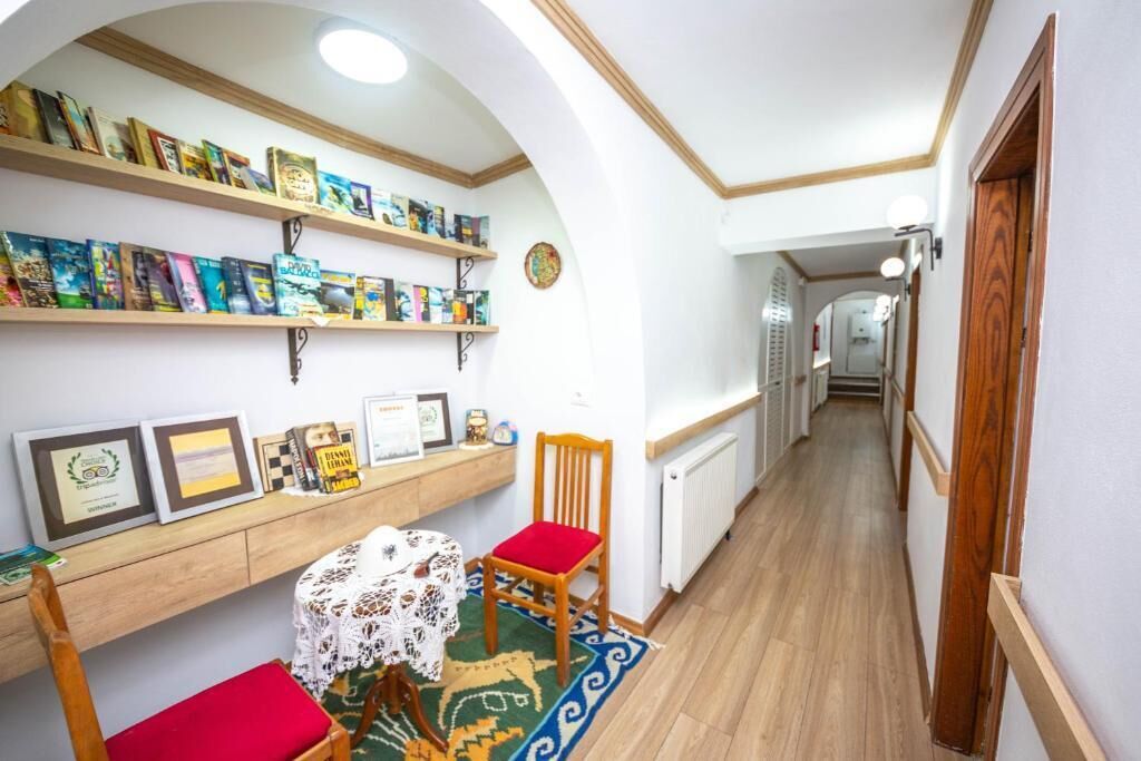 A narrow hallway with a wooden floor, a small table with a lace tablecloth, and a chair with a red cushion. Shelves above the table hold books and framed certificates.