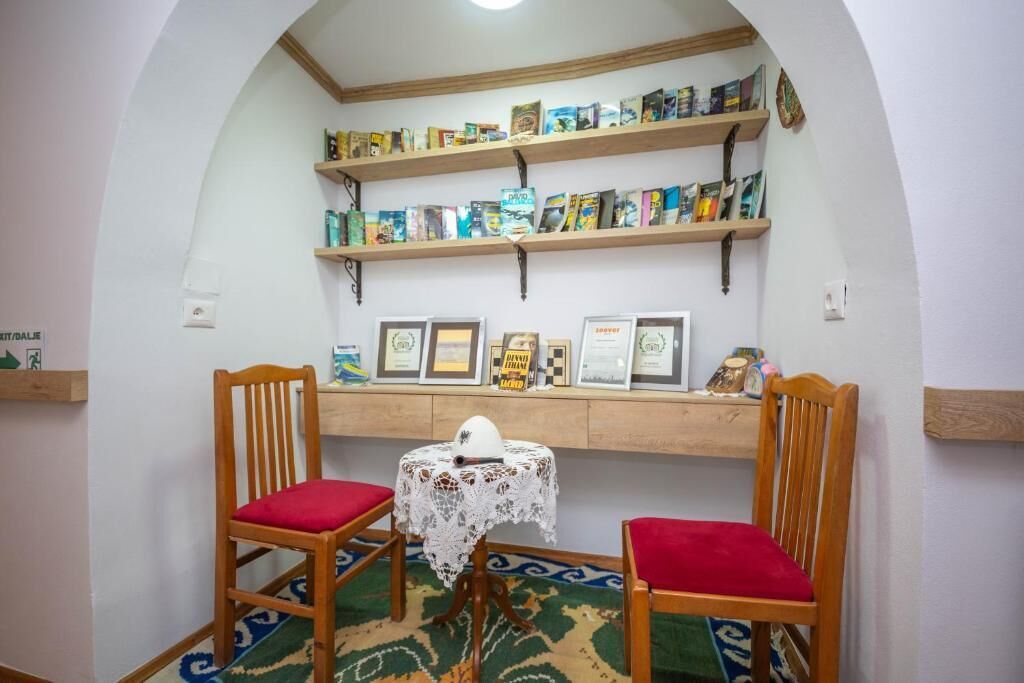 Een kamer met twee houten stoelen met rode kussens, een tafeltje met een kantkleed en daarboven boekenkasten met boeken en ingelijste objecten.
