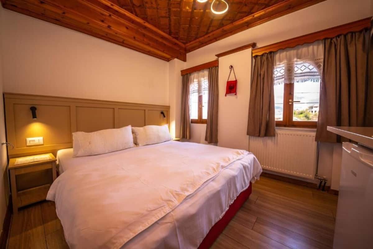 A bedroom with a wooden ceiling, a bed with white bedding, a nightstand, a window with curtains, and a radiator.