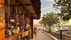 A seaside bar with wooden seating and hanging lights, overlooking a beach with umbrellas and lounge chairs. - Upgraded 1BR Paradise With Private Garden - Oceana (Dubai)