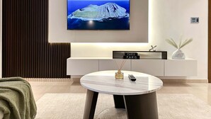 A modern living room with a flat-screen TV mounted on the wall, a round coffee table with a vase and a remote control, and a minimalist console table with a decorative item. - Upgraded 1BR Paradise With Private Garden - Oceana (Dubai)