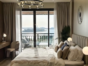 A bedroom with a bed, a desk, a chair, a balcony with a view of the sea, and a modern chandelier. - Fabulous 1 Bedroom Apartment at 5 Resort (Dubai)