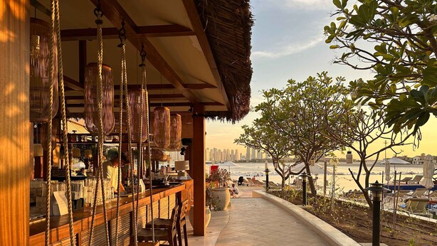 A seaside bar with wooden seating and hanging lights, overlooking a beach with umbrellas and lounge chairs. - Renoveted Marina View Apartment in Oceana (Dubai)