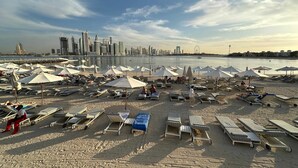 A beach with rows of sun loungers and umbrellas, a city skyline in the background, and a calm sea. - Renoveted Marina View Apartment in Oceana (Dubai)