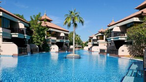 A large swimming pool with a central island, surrounded by multi-story buildings with balconies and palm trees. - Stunning Renovated 1BR Apartment at 5 Star Resort (Dubai)