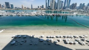 Una marina con barche ormeggiate, una spiaggia sabbiosa con ombrelloni e lettini, e uno skyline cittadino con moderni grattacieli sullo sfondo.
