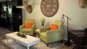Ein Wohnzimmer mit Deckenventilator, einer großen Wanduhr, zwei Sesseln mit orangefarbenen Kissen, einem Couchtisch und einem Teppich.