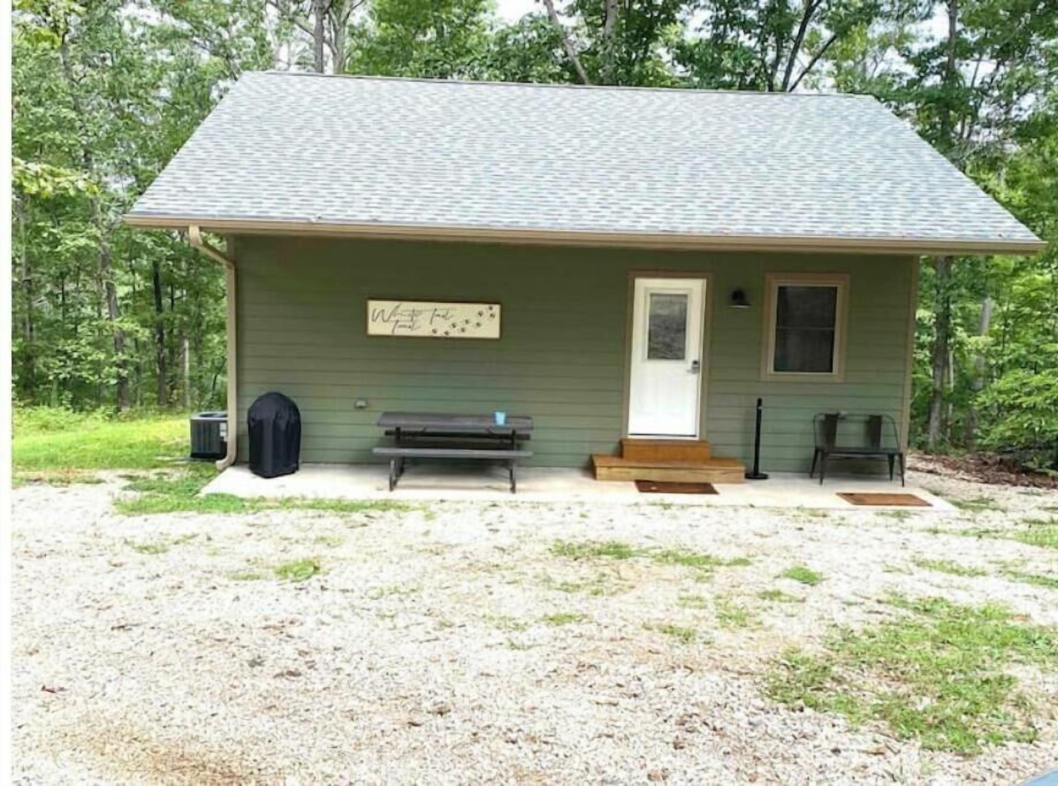 A small cabin with a green exterior, a white door, a bench, and a picnic table outside.