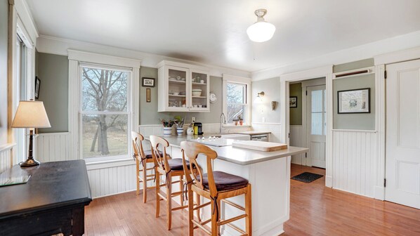 A kitchen with a central island, wooden chairs, a dining table, and a window with a view of trees. - Cozy Sag Harbor home close to town and beaches, great for families (Sag Harbor)