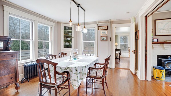 A dining room with a table set for four, surrounded by wooden chairs. There are large windows on one side, a bookshelf, and a fireplace with a map on the wall. - Cozy Sag Harbor home close to town and beaches, great for families (Sag Harbor)