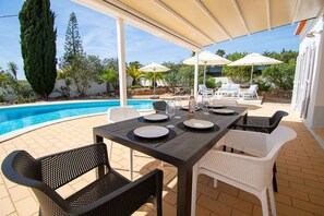 Ein Essbereich im Freien mit Pool, weißen Korbstühlen und Sonnenschirmen.