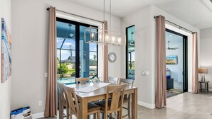 Un'area pranzo moderna con tavolo e sedie in legno, ampie finestre con tende, un orologio ben visibile e vista su una piscina e vegetazione all'esterno.