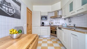 Une cuisine moderne avec des meubles blancs, un four à micro-ondes, un four et un évier. On y trouve une table en bois, un saladier rempli de fruits et une chaise.
