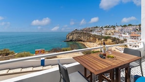 Un balcon avec une table en bois, des chaises blanches et une vue sur la mer et une ville côtière.