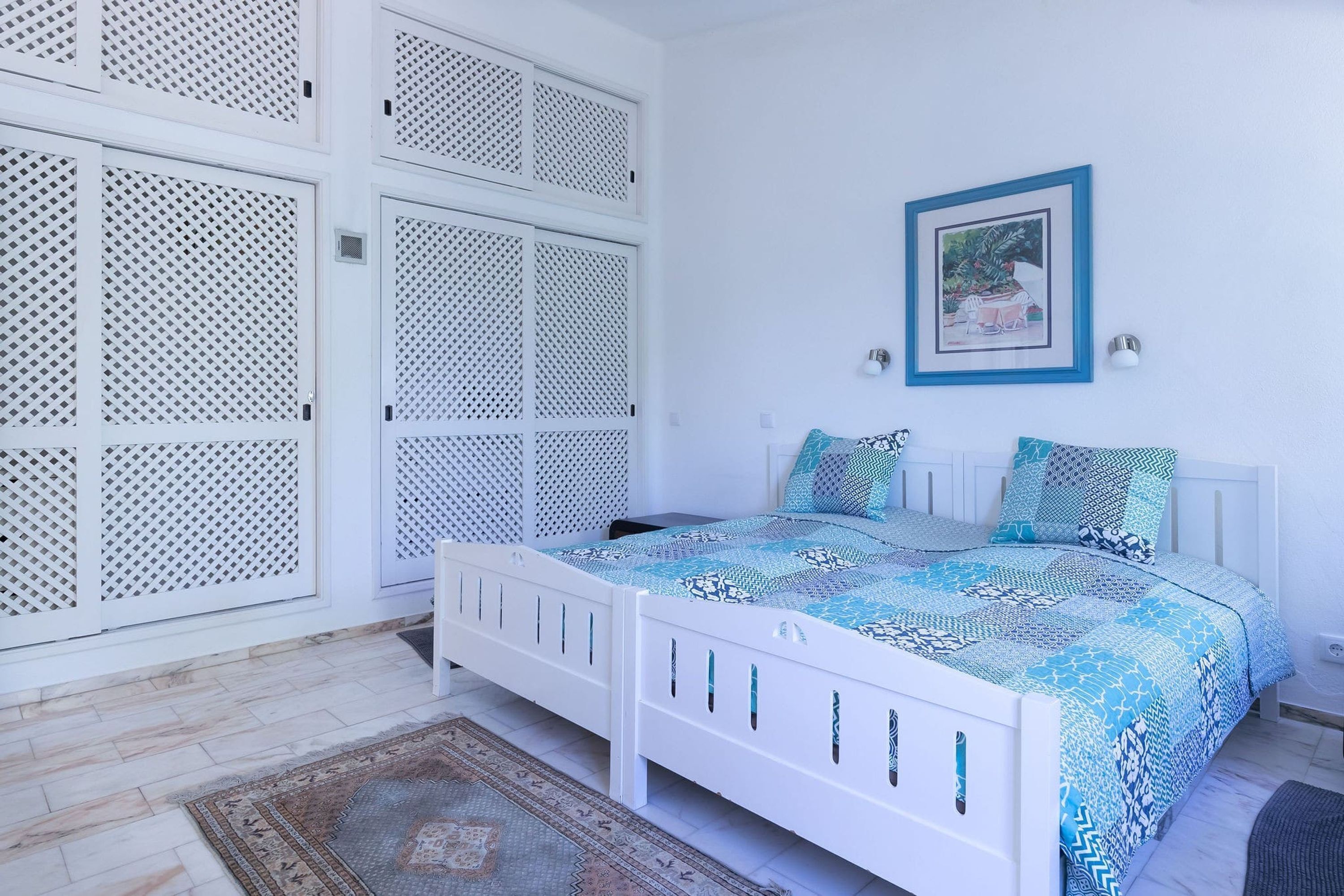 A bedroom with a white bed, patterned bedding, a framed picture on the wall, and a patterned rug.