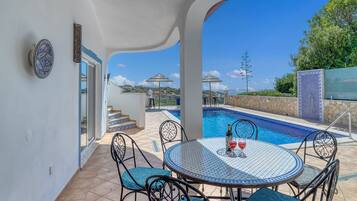A poolside area with a round table, chairs, and a glass of red wine.
