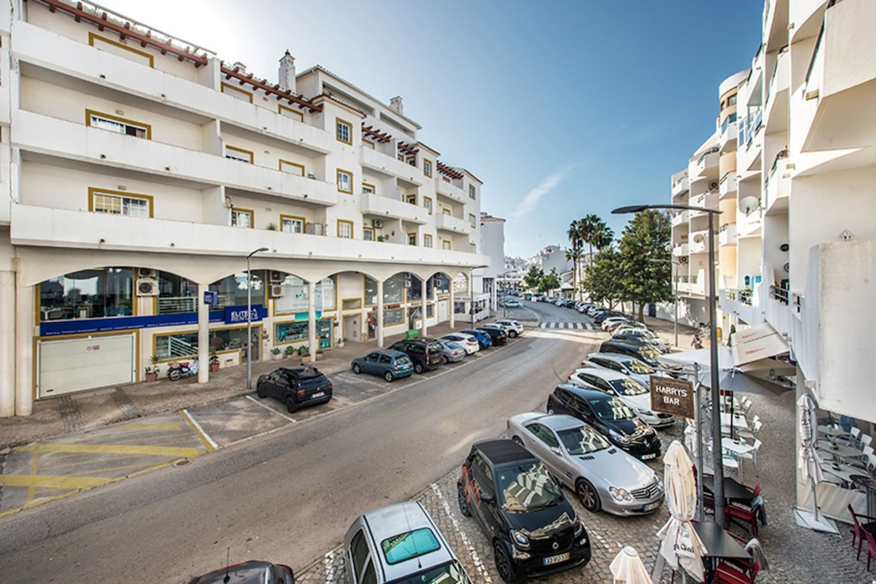 A street view with a row of parked cars, a building with multiple floors, and a restaurant named "HARRY'S BAR" on the right.
