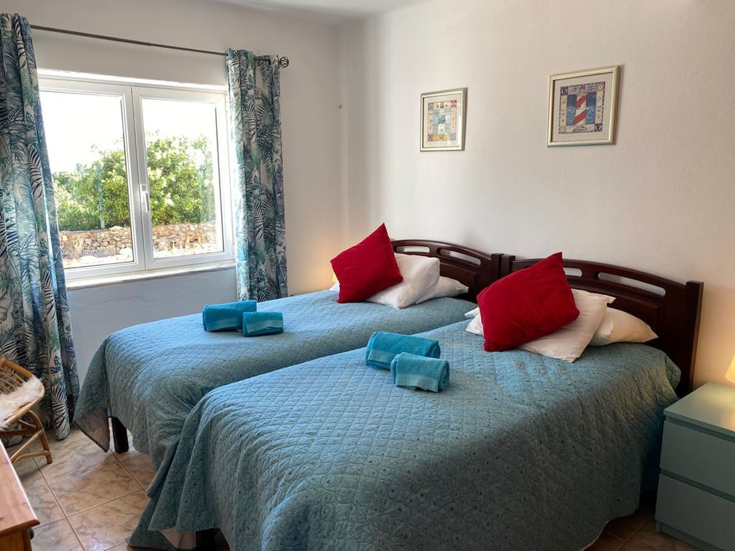Una habitación con dos camas, cada una con cubrecama azul y almohadas rojas, una ventana con cortinas y dos cuadros enmarcados en la pared.