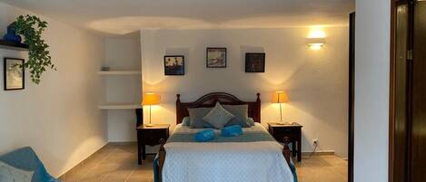 A bedroom with a bed, two bedside tables, a nightstand, a sofa, and a wall-mounted shelf with a plant.