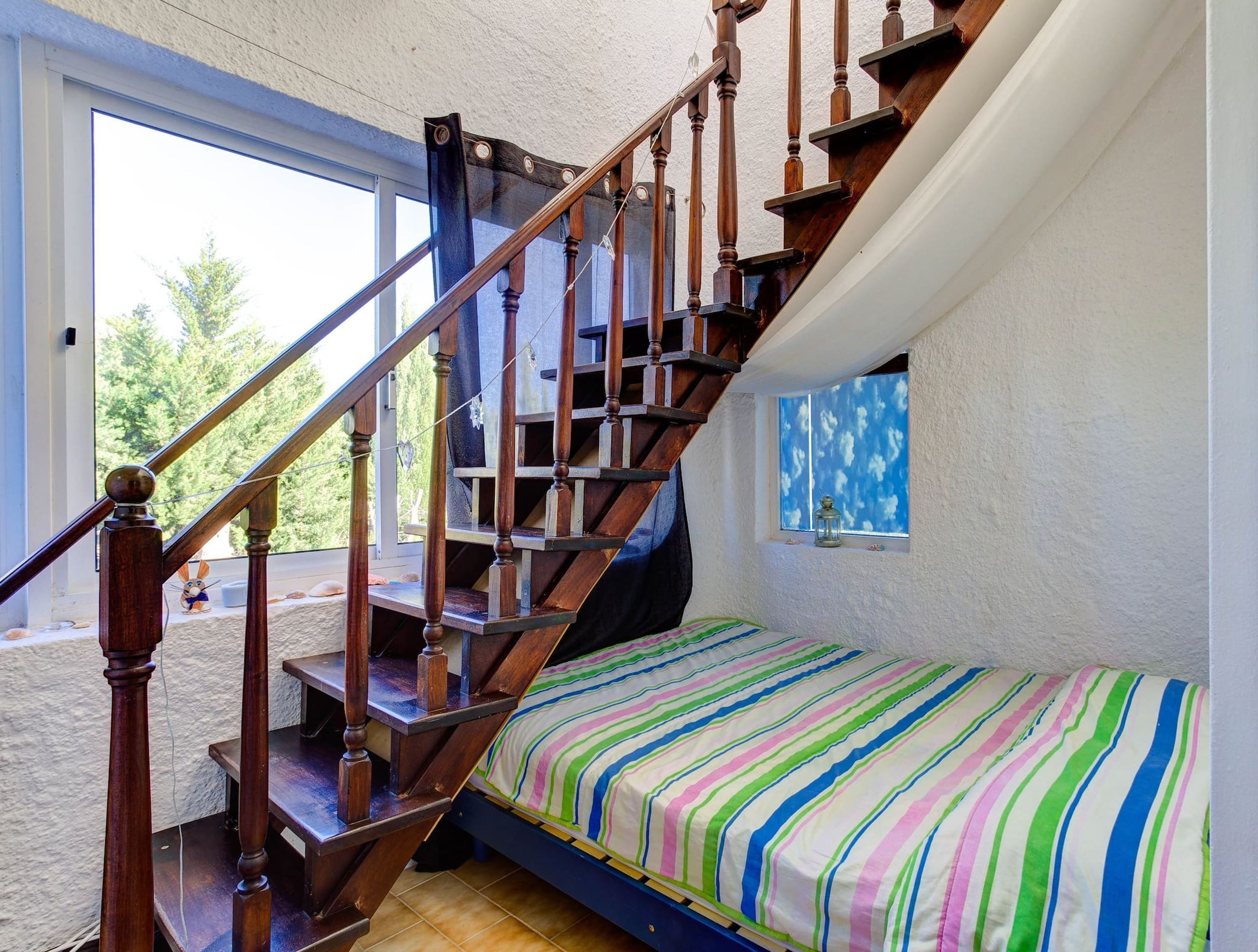 Een slaapkamer met een houten trap, een kleurrijk gestreept bedovertrek en een raam met uitzicht op bomen.