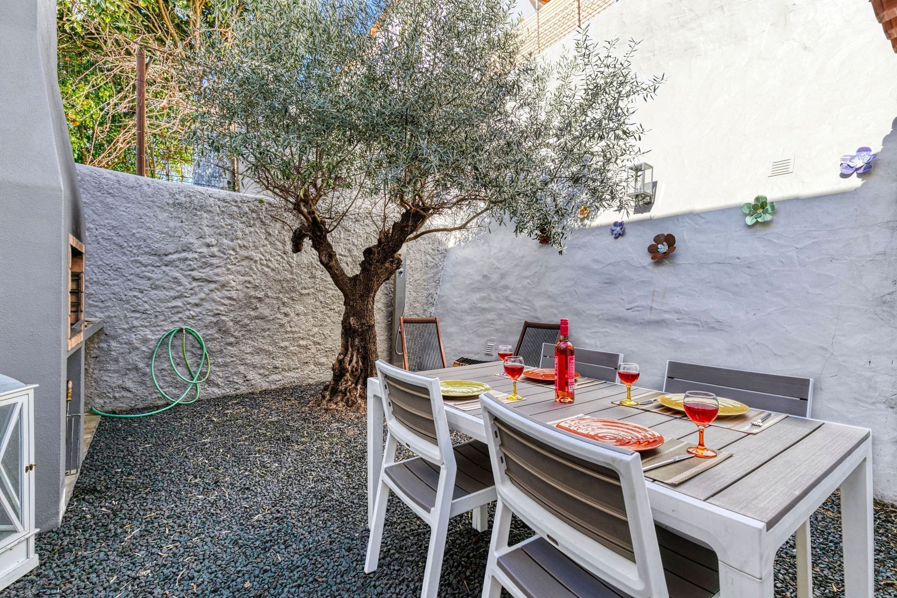 A patio with a table set for two, surrounded by white chairs, a tree, and a white wall with flower decorations.