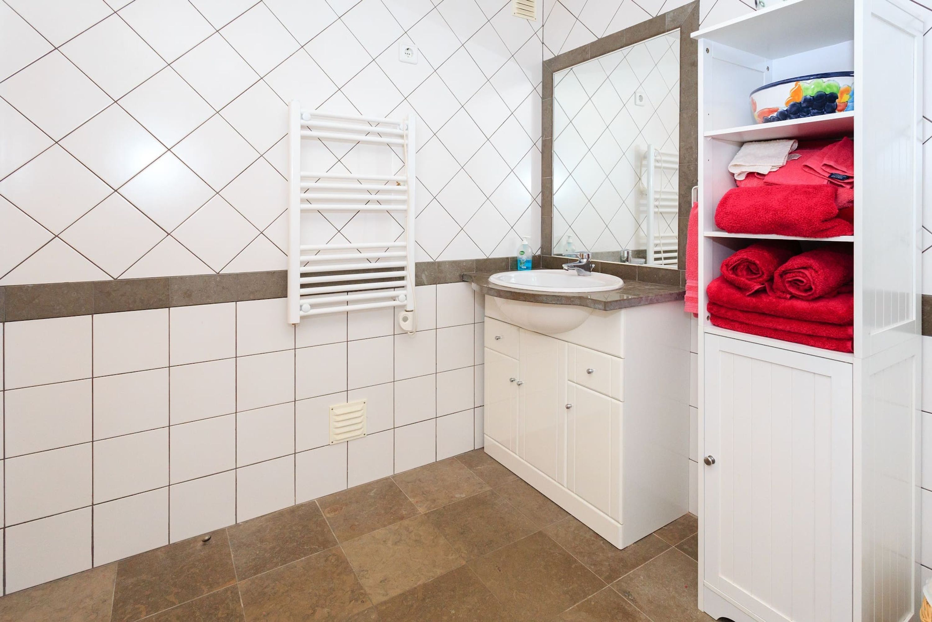 Une salle de bain avec des murs carrelés de carreaux blancs, un sol carrelé de carreaux bruns, un lavabo blanc, un miroir et un porte-serviettes.