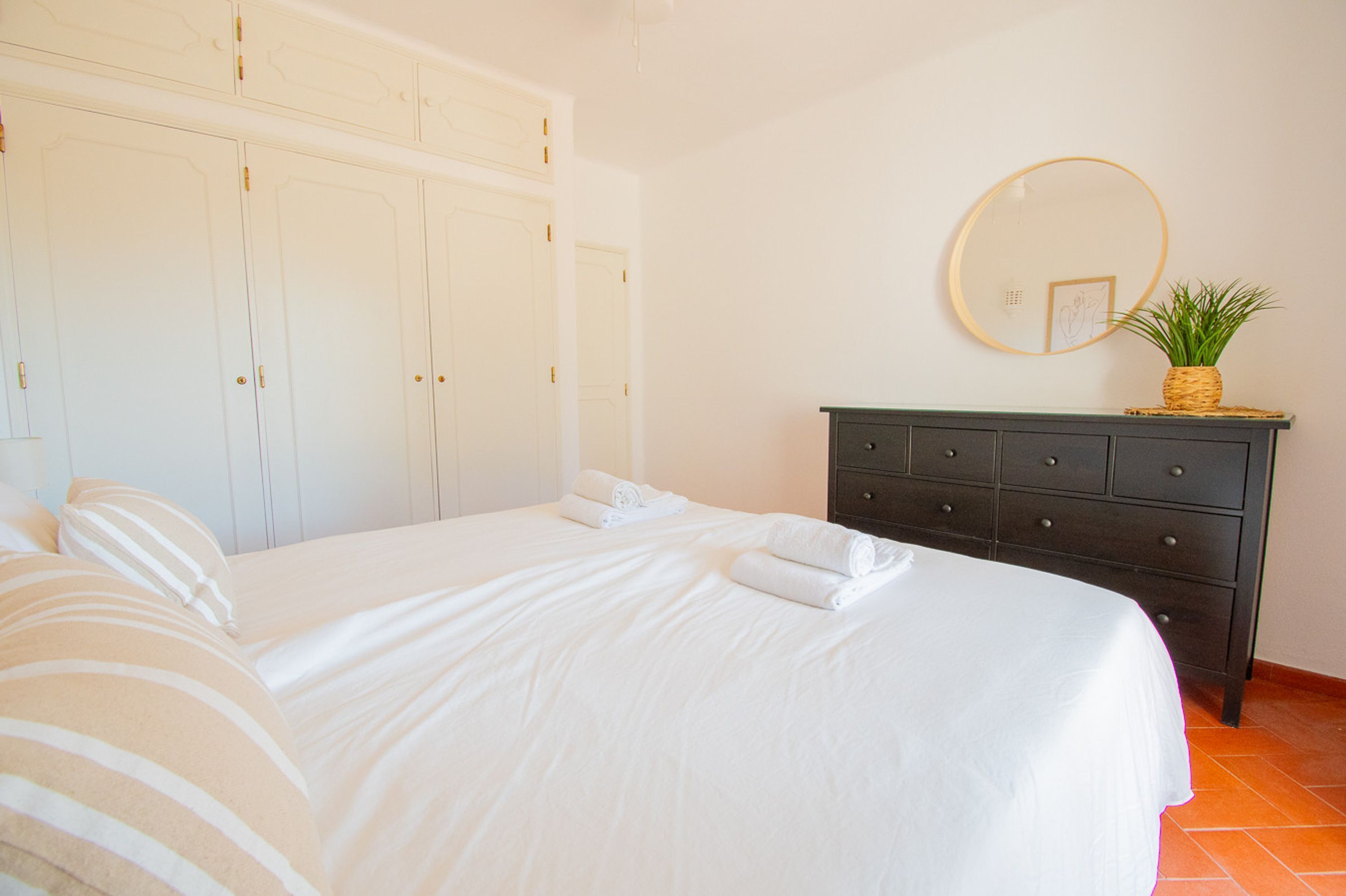 A bedroom with a large bed, white curtains, a dark dresser, and a round mirror on the wall.
