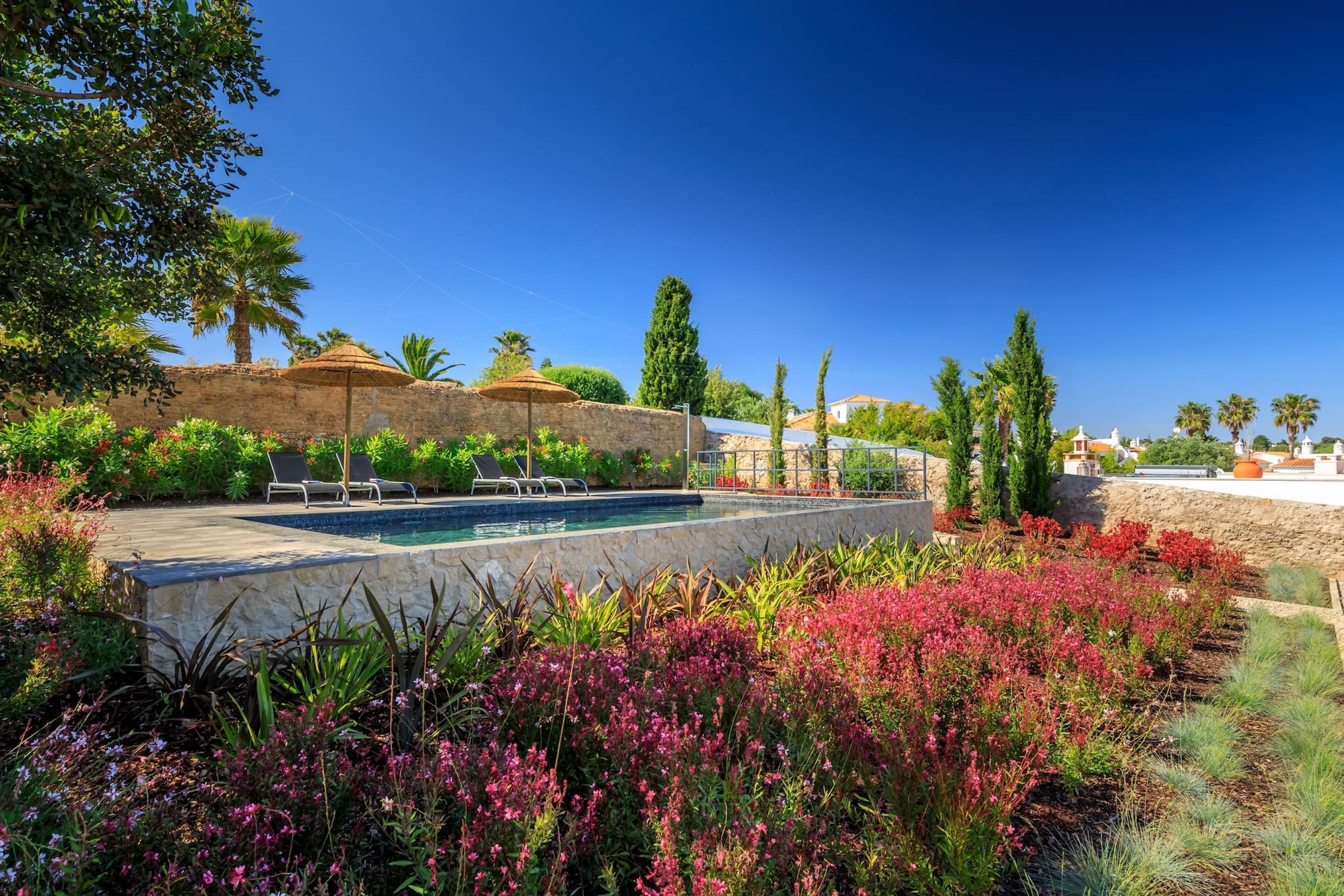 Et poolområde med liggestole og parasoller, omgivet af frodig vegetation og blomstrende planter.