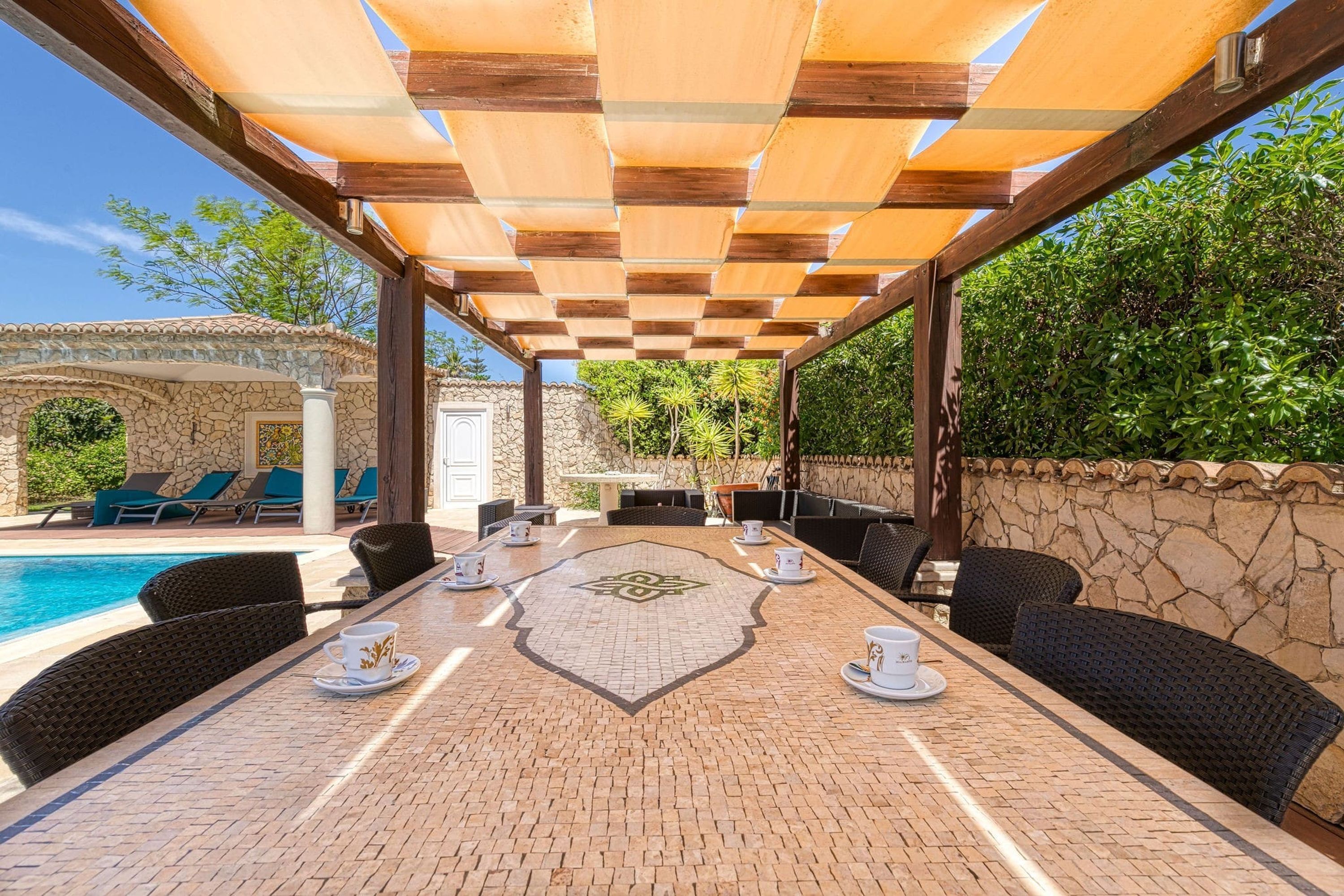 An outdoor patio with a long table, chairs, and a pool in the background.