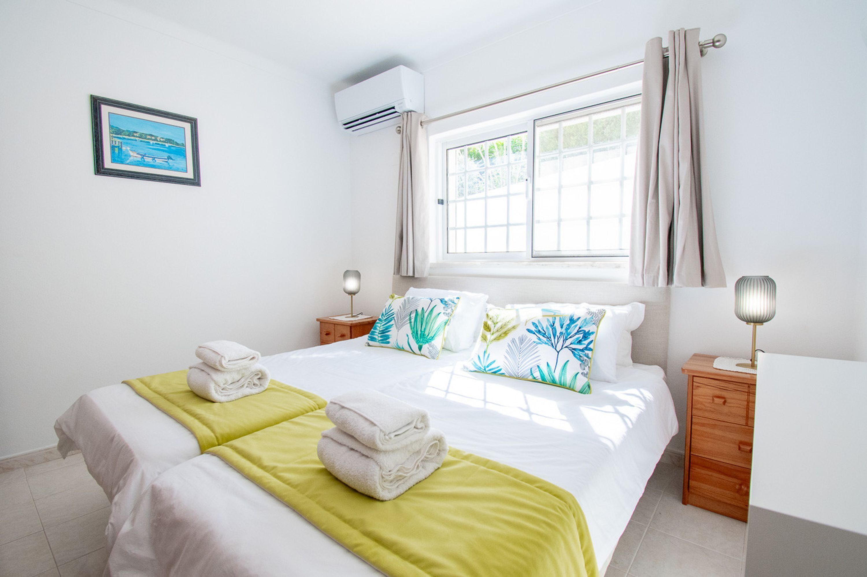 A hotel room with a bed, two bedside tables, a nightstand, a window with curtains, an air conditioning unit, and a framed picture on the wall.