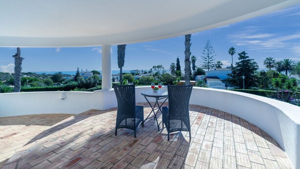 Un patio esterno coperto con tavolo e sedie, affacciato su un panorama pittoresco con alberi e vista sul mare.