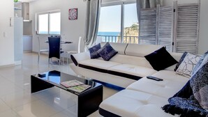 Un salon moderne avec un canapé blanc, une table basse en verre et une vue sur la mer grâce à de grandes fenêtres.