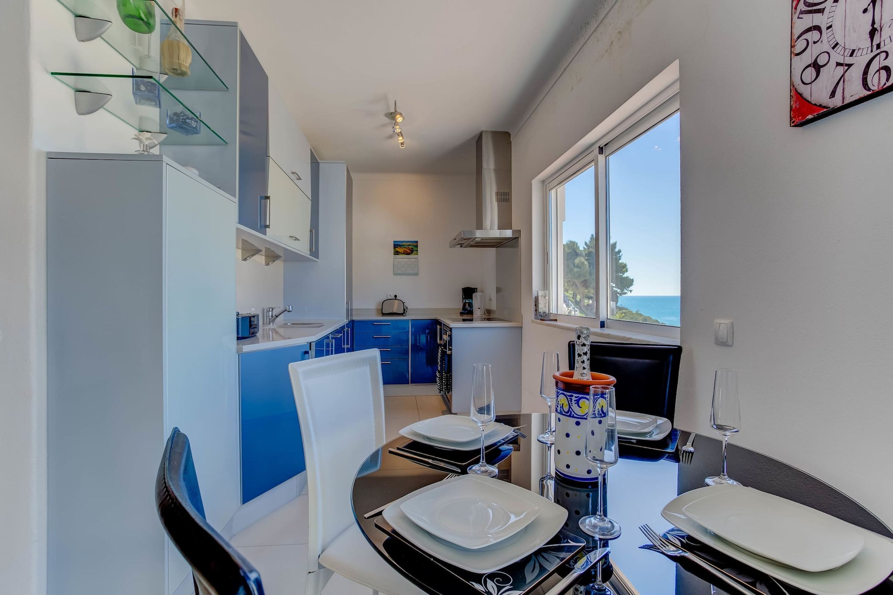 Une cuisine moderne avec des meubles bleus, un coin repas avec une table dressée pour deux personnes, et une grande fenêtre offrant une vue sur la mer.
