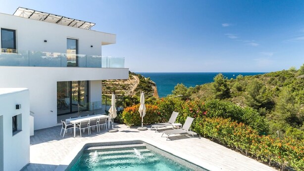 Una casa moderna de dos pisos con piscina, mobiliario exterior y vista al mar.